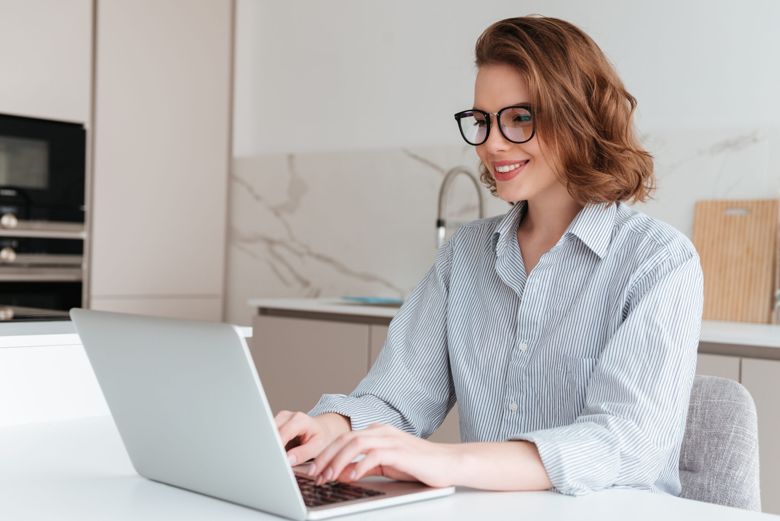 elegant-smiling-woman-glasses-striped-shirt-using-laptop-computer-while-siting-table-kitchen-scaled