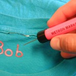 Image of fluorescent pink paint in Metron Marker being held over a teal shirt with the name BOB that was written in the pink paint.